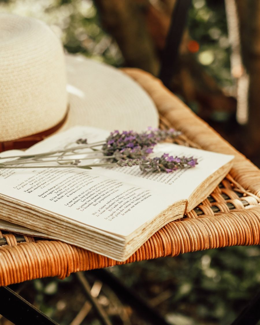 An open book with a sprig of lavender beside a straw hat on a wicker chair
